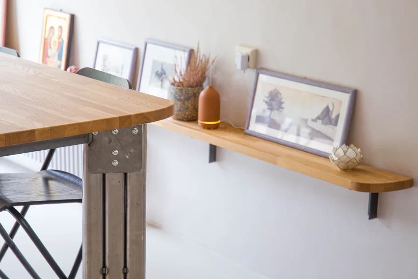 Corner of an oak bar table beside a floating shelf styled with framed prints and a ceramic diffuser