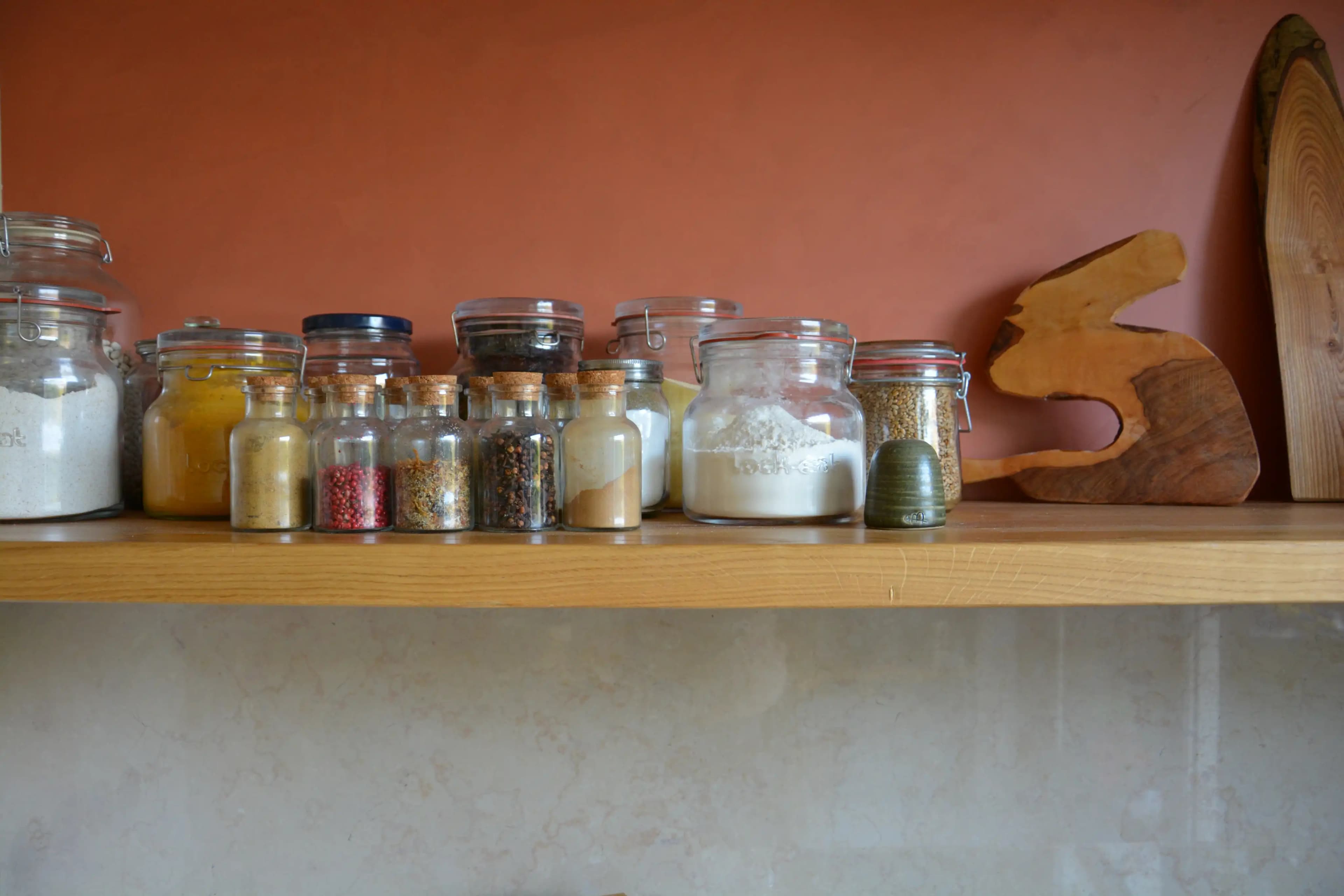 Glass spice jars lined up on a wooden shelf against a terracotta wall